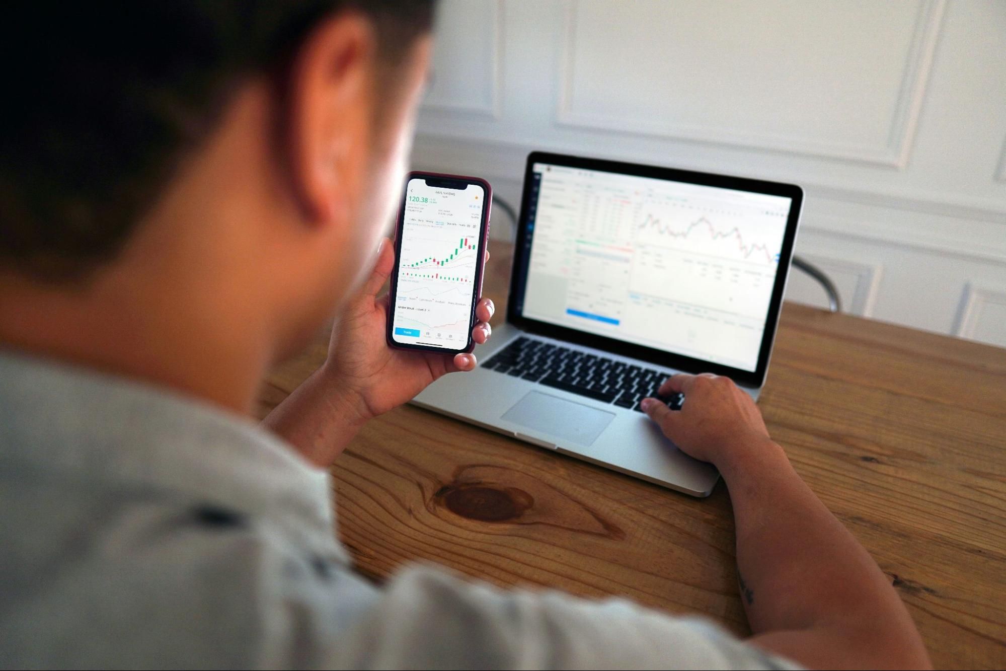 Man looking at financial charts displayed on his smartphone and laptop screen.
