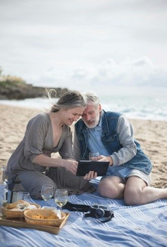 An elderly man and woman sitting on a blanket having a picnic by the seaside.